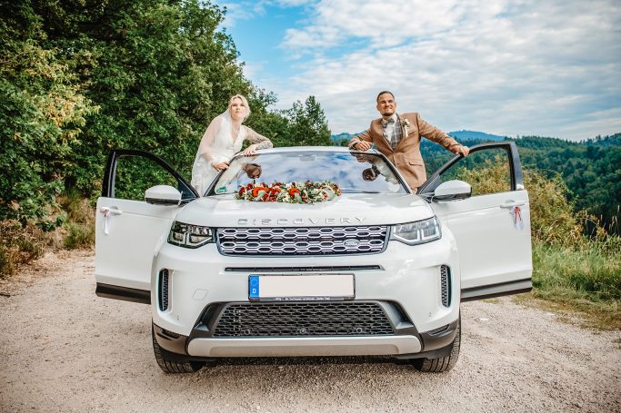 Verliebtes Paar steht nebeneinander an einem Auto, Blick in die Kamera